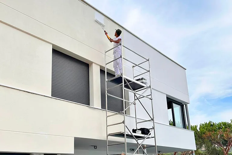 Pintor con rodillo pintando una fachada blanca desde un andamio